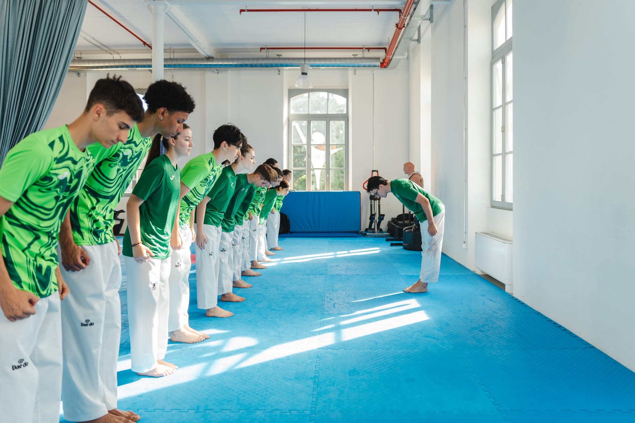 Gruppo di ragazzi in maglietta verde Summit Taekwondo che si inchinano all'istruttore nella palestra luminosa con grandi finestre